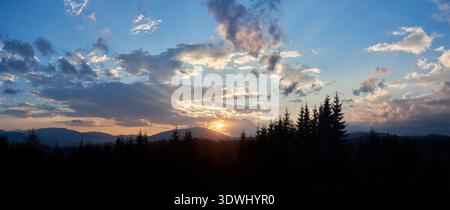 Sun sets behind range of distant mountains, casting golden rays through clouds. Sky, painted with shades of blue, pink, and gold, in tranquil evening. Silhouetted pine trees add depth to landscape. Stock Photo
