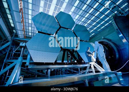 From 2010, two engineers in cleanroom suits meticulously work on segments of the James Webb Space Telescope's primary mirror at NASA's Goddard Space Flight Center. The hexagonal mirror segments, crucial for the telescope's deep space observations, are seen under bright, specialized lighting, highlighting the precision and scale of the project. Stock Photo