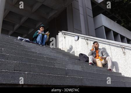 Diverse college-aged male students sitting on campus steps writing notebook holding phone, bag, cup Stock Photo
