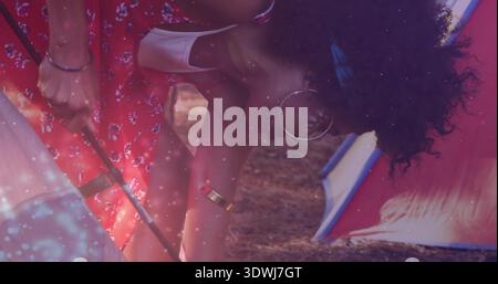 Adjusting woman with hoop earrings assembling tent on pine-needle, pal holding pole in red dress Stock Photo