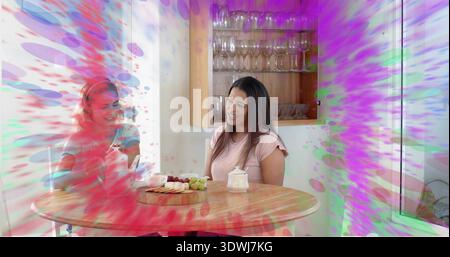 Chatting women wearing blue and pink tops sitting at table in home kitchen, sharing serving board Stock Photo