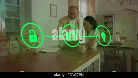 Reviewing senior couple wearing cardigans using laptop at dining area, with smart-home icons, mugs Stock Photo