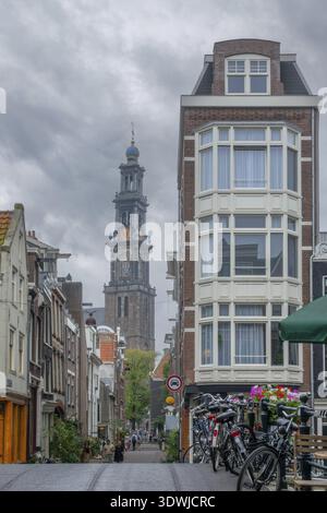 Netherlands. Cloudy summer day in Amsterdam. People and bicycles on a narrow street with typical houses in the city center. Old cathedral with bell to Stock Photo
