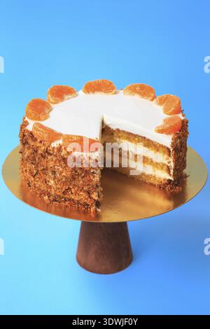 Sliced Birthday cake on the wooden cake stand. Beautiful orange sponge cake with whipped cream decorated with tangerine slices. Blue background. Copy Stock Photo