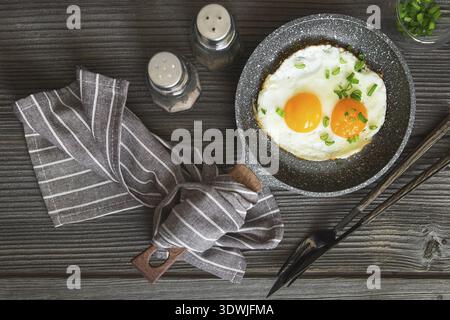Fried eggs with green chives chopped for breakfast. Two fried eggs in a small pan on the dark wooden background, top view. Flat lay. Rustic style Stock Photo