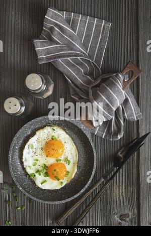 Fried eggs with green chives chopped for breakfast. Two fried eggs in a small pan on the dark wooden background, top view. Flat lay. Rustic style Stock Photo