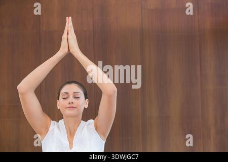 Woman raising arms up pressing palms together meditating by wood wall wearing white sleeveless top. Yoga, mindfulness, serenity, calm, balance, studio Stock Photo