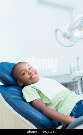 African American boy reclining in blue dental chair at operatory, wearing green T-shirt, white bib. Pediatric, clinic, calm, comfort, reassurance, ove Stock Photo