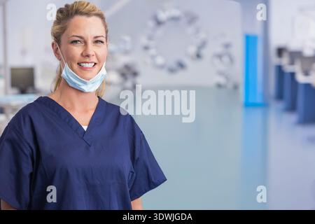 Mid adult woman smiling, posing in operating room in navy scrubs with mask lowered, copy space. Clinician, hospital, surgical, healthcare, cheerful, m Stock Photo