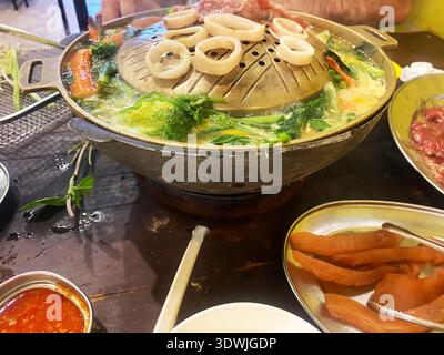Hot pot meal featuring fresh vegetables, seafood, and meat on a table in a restaurant setting with a steaming pot and various dipping sauces Stock Photo
