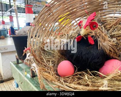 Chicken mannequin with decorative features and pink eggs nestled in a woven basket on a market table, surrounded by a vibrant market setting Stock Photo