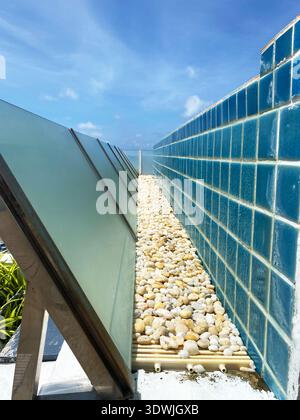 Decorative gravel drainage area with smooth white stones between glass panels and blue tiled wall under clear sky in a modern architectural setting Stock Photo