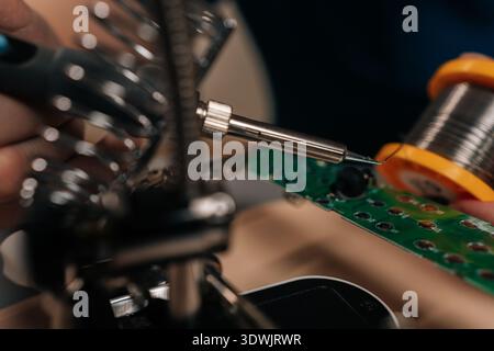 Close-up of technician soldering electronic circuit board using magnifier tool for precise repair and assembly work in modern engineering and Stock Photo