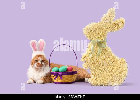 Cute cat in bunny ears with Easter basket and decorative rabbit on lilac background Stock Photo