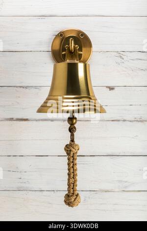Vintage brass bell used on ships, with knotted rope, hanging on hook mounted to rustic wooden wall Stock Photo