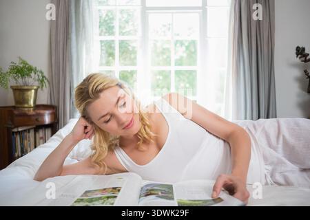 Adult woman reclining on bed in bedroom, reading magazine by paned window and bedside plant. Female, relaxation, sunlight, cozy, minimalist, interior, Stock Photo