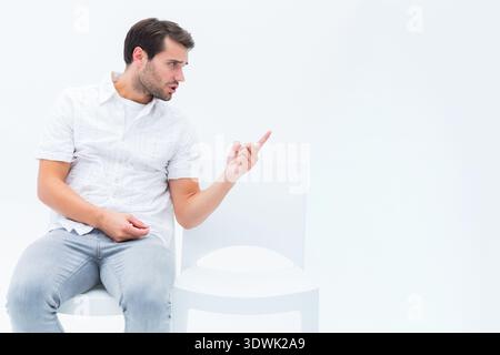 Man sitting on white chair, turning and pointing at empty white chair in studio, copy space. Male, solo, minimal, highkey, clean, gesture, direction Stock Photo