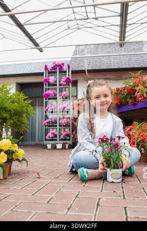 Child girl sitting cross-legged on brick floor in greenhouse holding white pot with pink flowers. Kid, greenery, plants, glasshouse, sunlit, nursery, Stock Photo