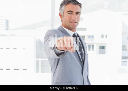 Man in 40s standing in bright office by floor-to-ceiling windows, wearing light gray suit, pointing. Executive, professional, corporate, confident, le Stock Photo
