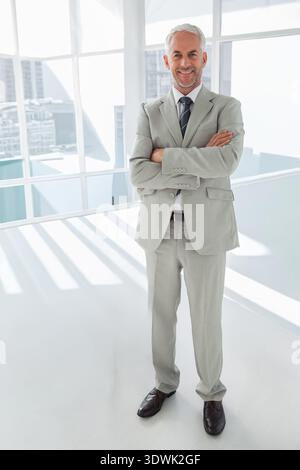 Senior man wearing suit, tie standing near glass wall in office, sunlight casting shadows on floor. Executive, workspace, modern, minimal, urban, skyl Stock Photo