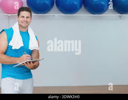 Adult male standing left in gym wearing turquoise-top, writing on clipboard near balls, copy space. Fitness, studio, trainer, notebook, ball, rack, pi Stock Photo