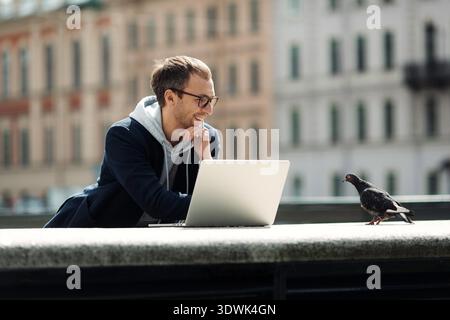 Millennial professional work outside using laptop computer and 5g internet connection. Successful smart casual web developer, designer man happy with freelance occupation and remote workspace in city || Model released Stock Photo