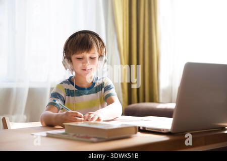 child boy in earphones is using a laptop and study online at home. homeschooling, distant learning || Model released Stock Photo