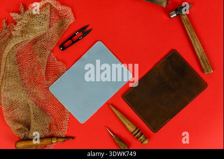 Two leather pieces, one blue and one brown, surrounded by crafting tools and burlap on a vibrant red background Stock Photo