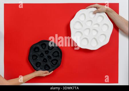 Two hands, one holding a black ceramic plate and the other a white ceramic plate, positioned against a bright red background Stock Photo
