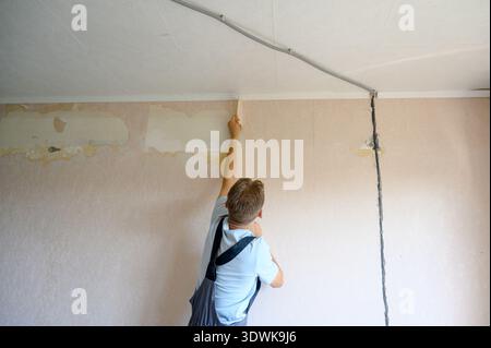 Male painter applies paint to the wall during a home renovation, with exposed electrical wires and unfinished sections of the wall in the background Stock Photo