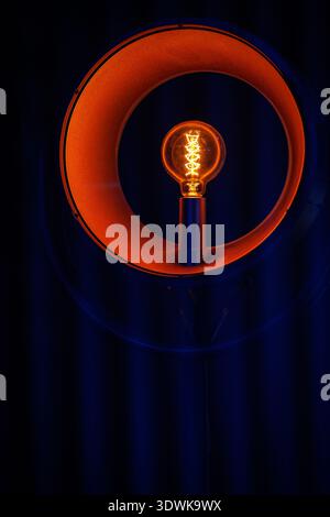 A close-up shot of a glowing filament light bulb set within a modern orange ring lamp. The background is dark blue with subtle vertical patterns. Stock Photo