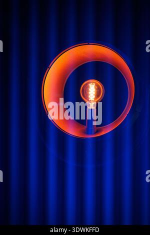 A single incandescent bulb glows orange within a round metallic frame. The background consists of vertical blue lines in shadow. Stock Photo