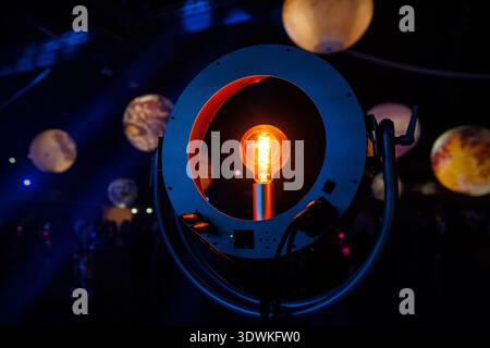 A glowing filament light bulb is centered within a round metal housing. The background features blue light and out-of-focus spherical decorations. Stock Photo