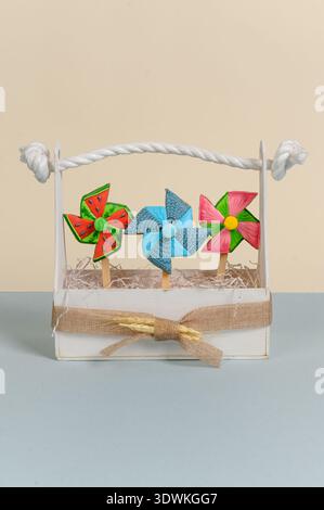Three vibrant pinwheels in a white basket with a rope handle, featuring colorful designs and placed on a light blue surface against a beige background Stock Photo