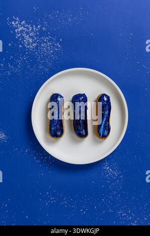 Three eclairs with blue glaze and sugar sprinkles arranged on a white plate, set against a vibrant blue background with a dusting of sugar Stock Photo