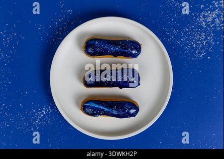 Three eclairs with blue glaze and decorative sprinkles arranged on a white plate, set against a vibrant blue background with sugar dust Stock Photo