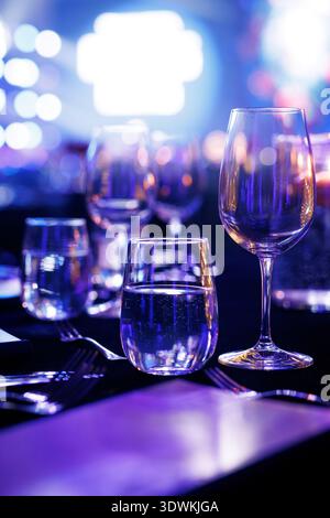 Clear glassware and silver forks sit on a dark table at a formal gala or conference, illuminated by vibrant blue and purple stage lights. Stock Photo