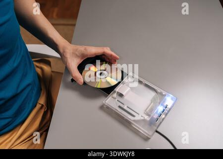 Top view of person inserting compact disc into an external optical disc drive connected to a laptop, transferring data for computer storage and media Stock Photo