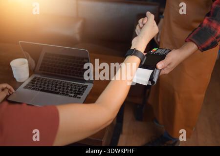 Close-up of a woman sitting in a coffee shop using a computer and making a contactless payment for a coffee with her smart watch Stock Photo