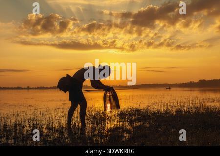 Side view silhouette of a fisherman standing in a river holding a fishing trap at sunrise, Thailand Stock Photo