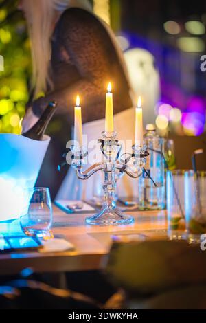 A table is set for dinner with a crystal candle holder in the center. Three candles burn brightly. Glasses are placed on the table as people prepare Stock Photo
