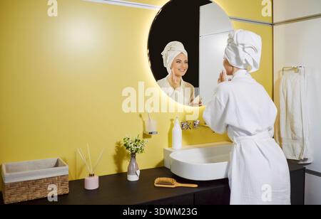 Happy beautiful woman looking in modern bathroom mirror, wearing white robe and towel wrapped Stock Photo