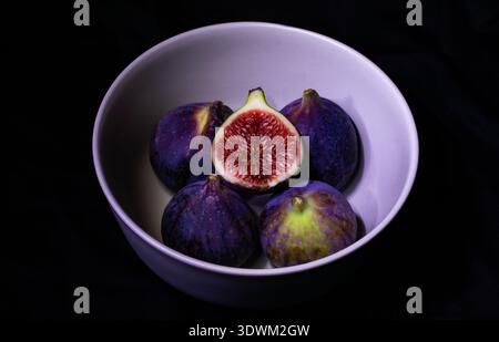 Ripe figs with contrasting purple and lime-green skin tones arranged around a cut fruit exposing juicy red flesh in a gray ceramic bowl on dark backdr Stock Photo