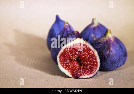 A halved fig with vivid red pulp and cream border leaning against three whole purple figs on a warm kraft paper surface with soft bokeh Stock Photo