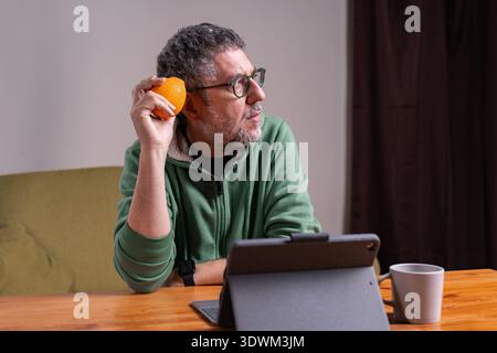 Senior man holding an orange and using a tablet at home during a modern morning routine. Stock Photo