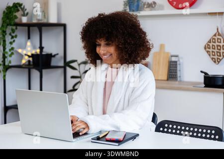 Smiling freelancer woman working from home in bathrobe using laptop Stock Photo