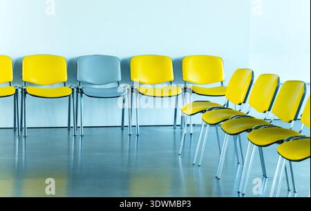 A line of unoccupied chairs, some bright yellow and one gray, are neatly arranged against a light blue wall, reflecting on the polished floor Stock Photo