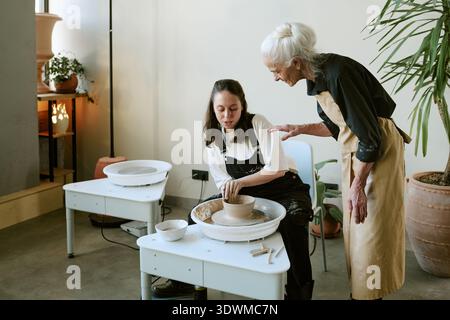 Senior Woman Instructing Young Adult Woman Shaping Clay Stock Photo