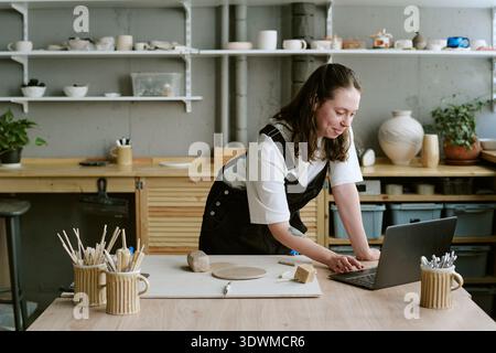 Young Adult Woman Sculpting Clay While Using Laptop in Pottery Studio Stock Photo