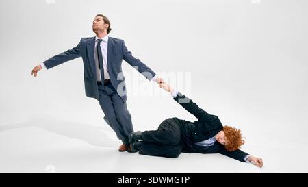 Businessman pulling colleague lying on floor in studio wearing dark suits on white background. Stock Photo